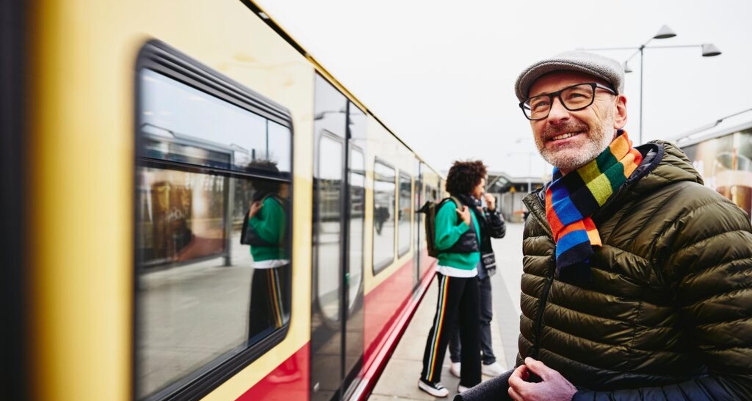 man in puffer jacket wearing rainbow scarf and beret smiling about to get on a train