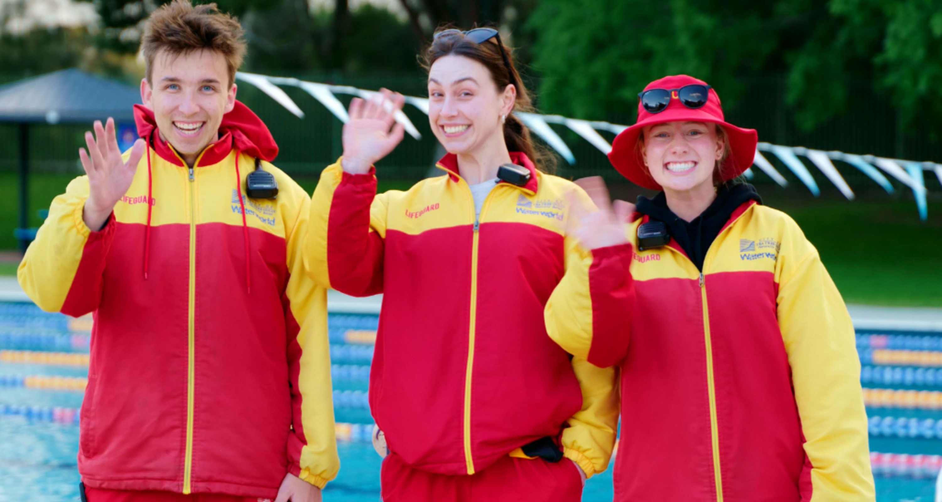 three lifeguards waving, one man, and two women in red and yellow jackets
