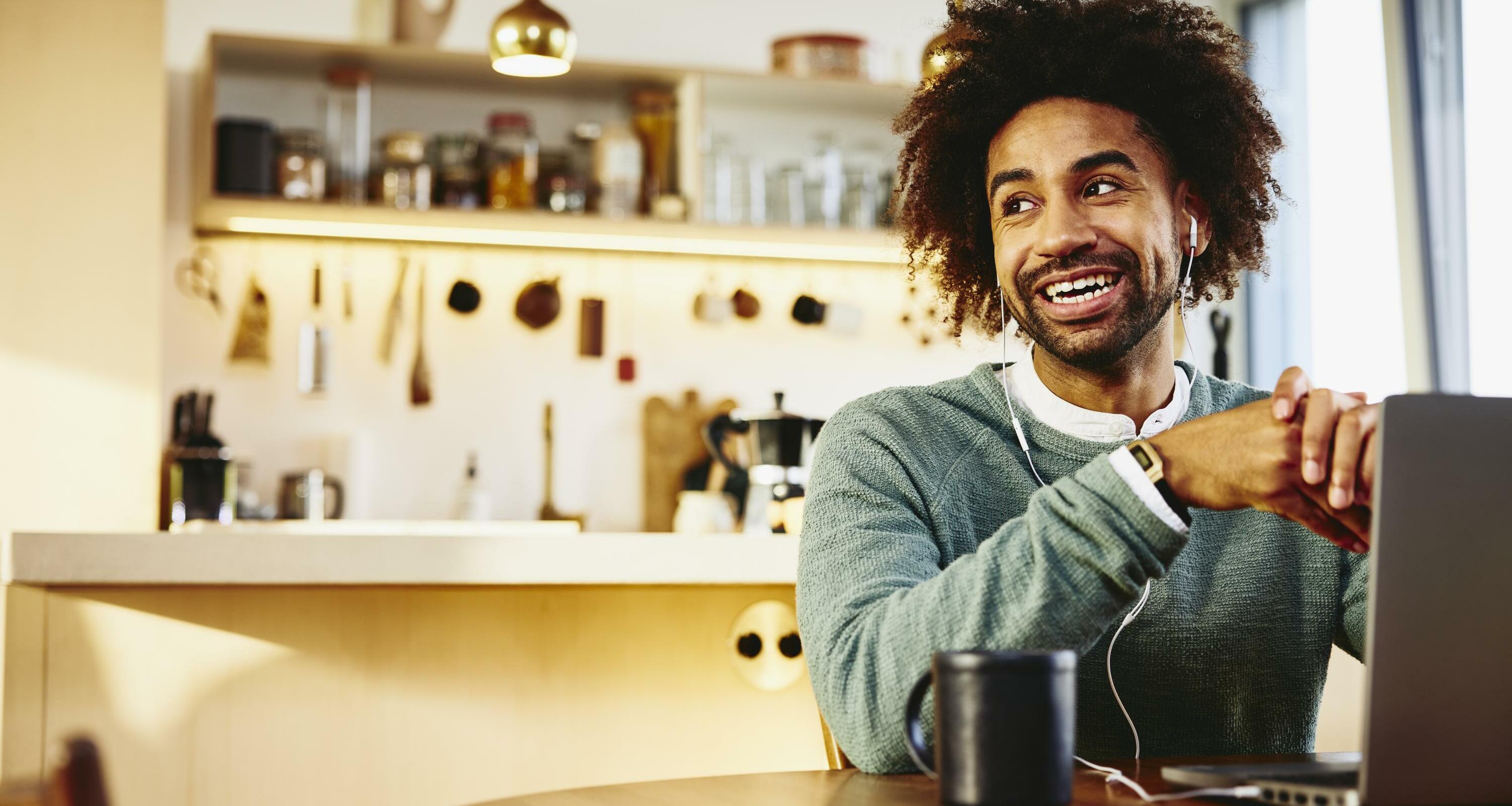 Smiling man sitting at his dining table with a drink and his laptop. Looking away. Kitchen in the background.