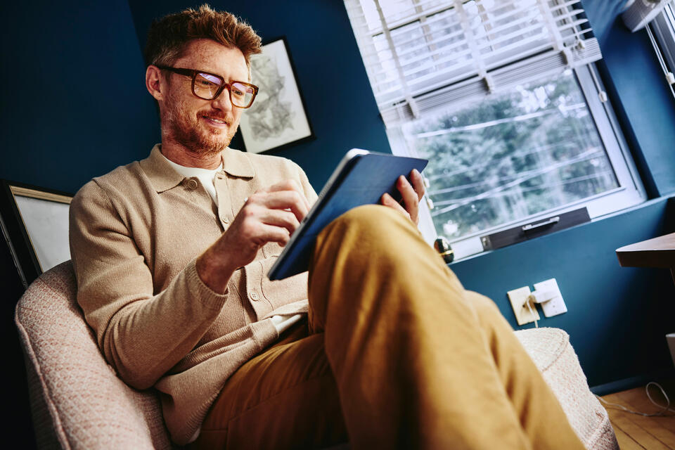an image of a man wearing glasses and sitting on a chair while using his tablet 