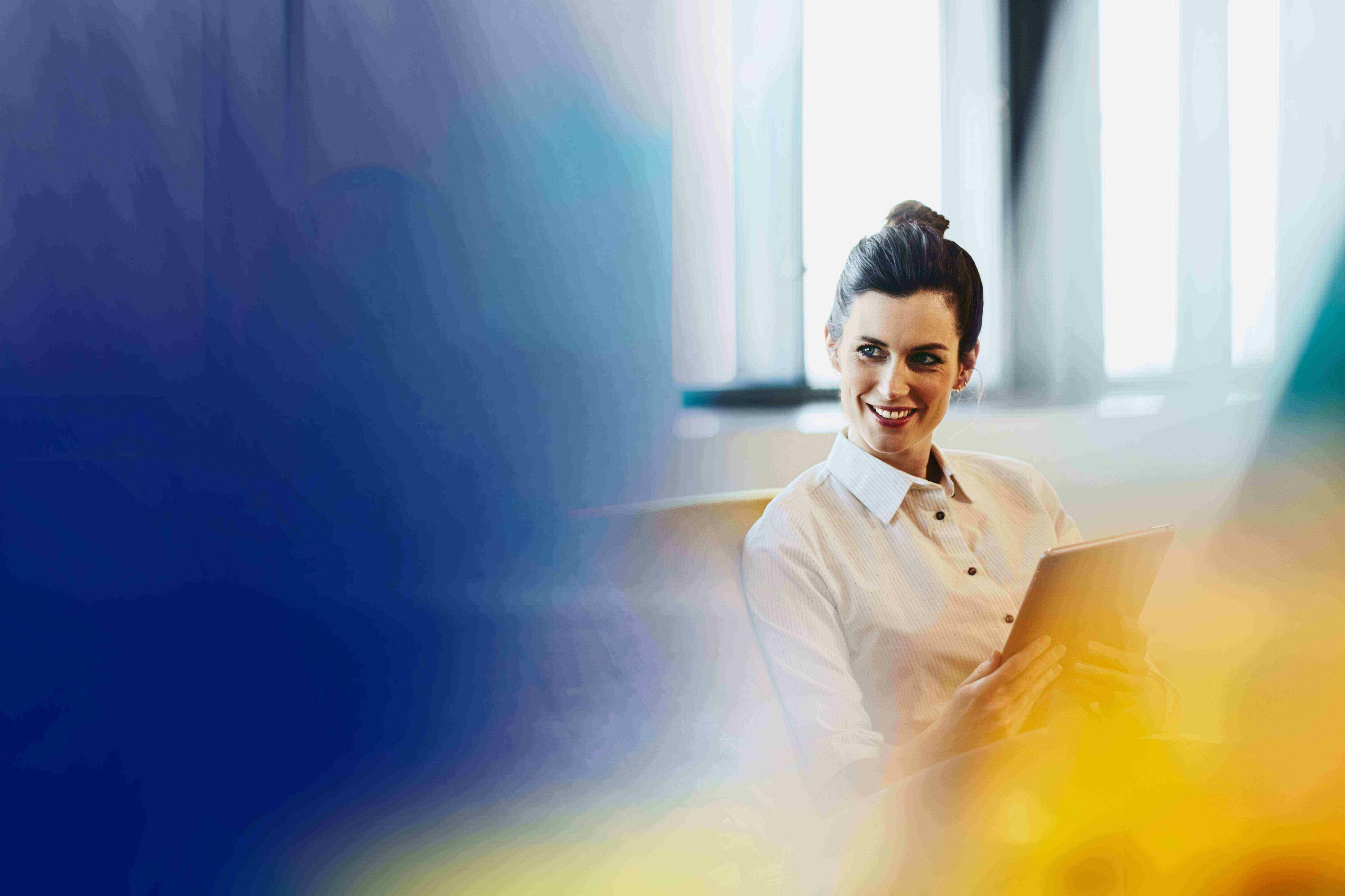 smiling woman holding an ipad sitting next to a window