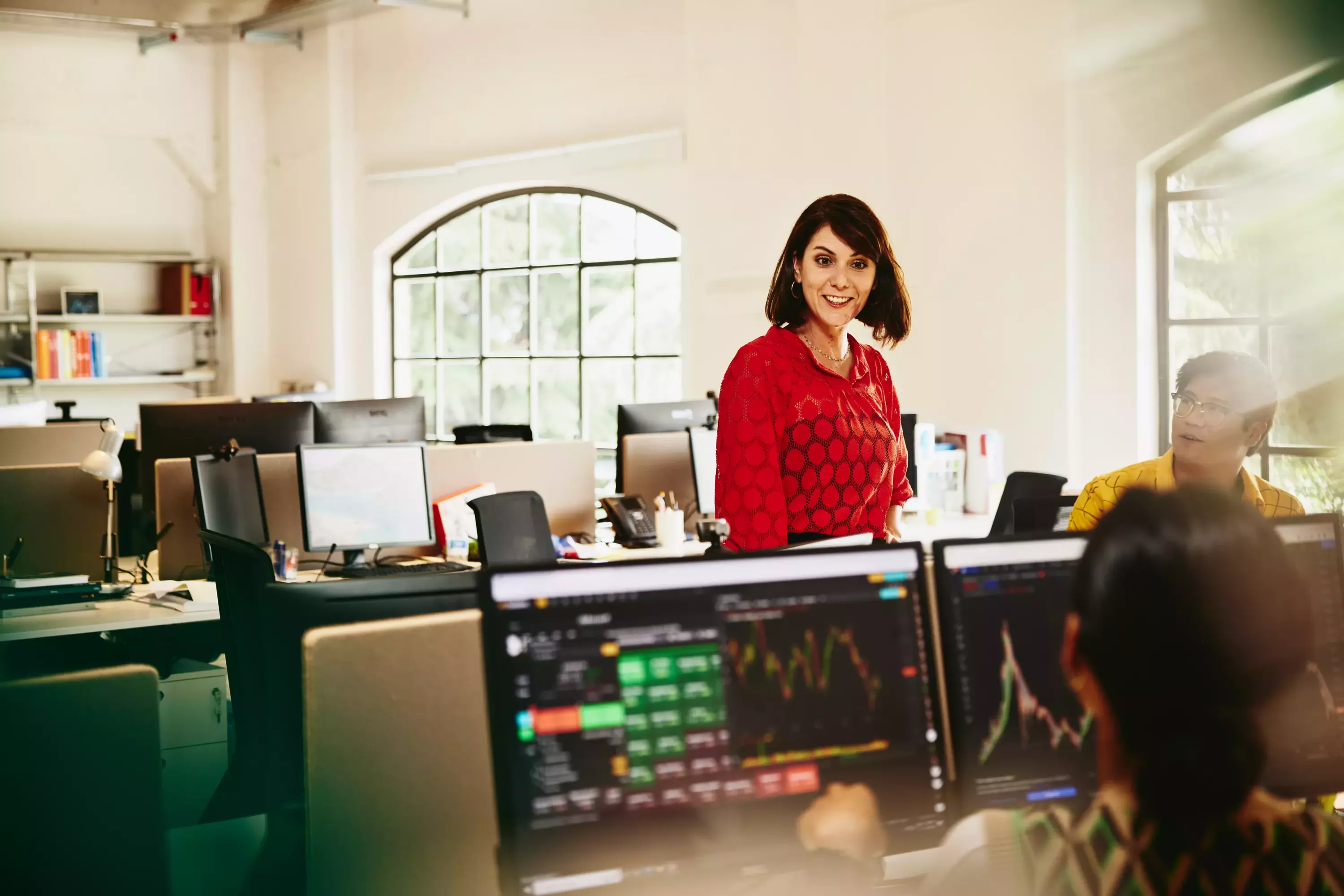 Smiling female talking to colleagues, standing between desks.

