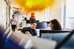 a man with headphones placed at his neck talking to a woman in the office  while there are people behind them