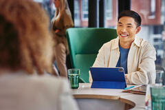 man in a cream jacket, blue top smiling in conversation with a colleague with a tablet in front of him