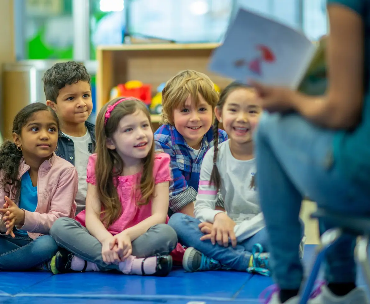 Group of children in a daycare centre. Their teacher is showing them a picture and they are smiling