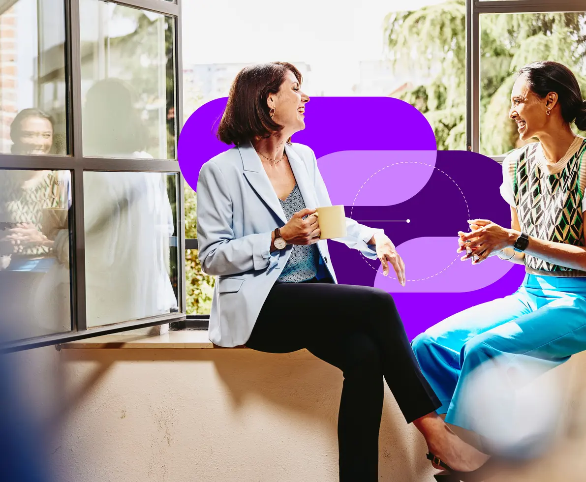 an image of two smiling female sitting on a window sill chatting and having coffee