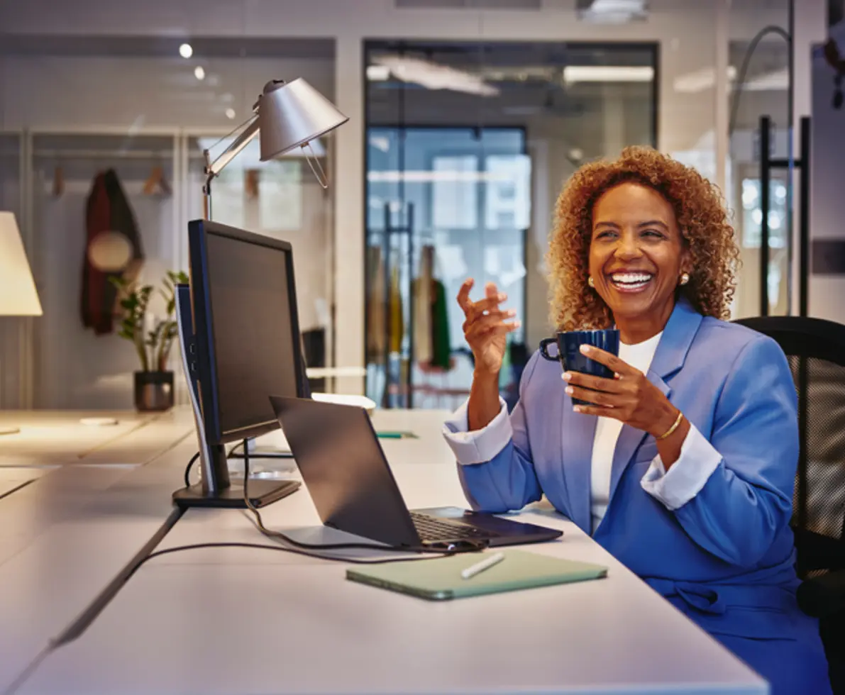 woman in blue blaser holding a mug smiling sitting at a desk 