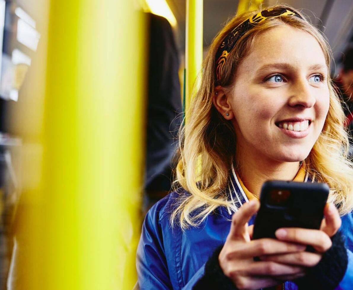 girl sitting on a bus with a phone in her hand she is smiling and looking out the window
