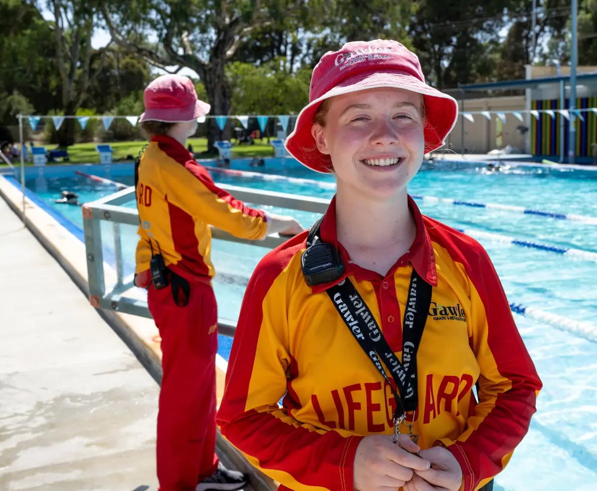 an image of two lifeguards standing outside a swimming pool. One is monitoring the pool and the other is facing the camera smiling