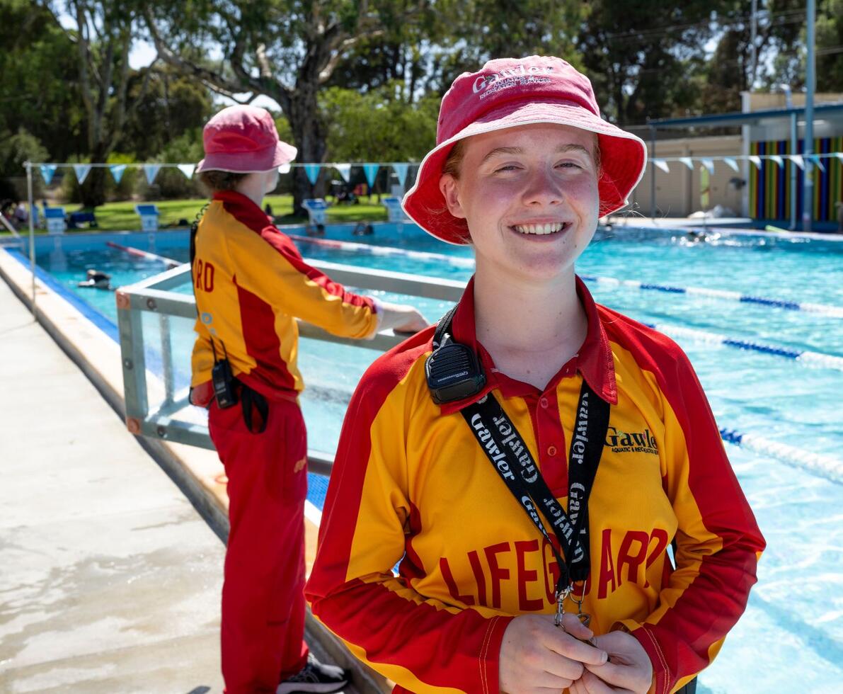 an image of two lifeguards standing outside a swimming pool. One is monitoring the pool and the other is facing the camera smiling
