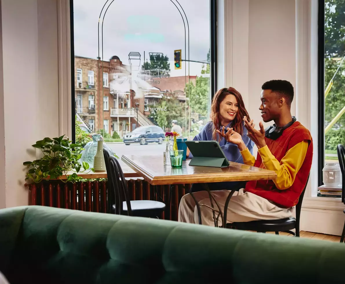 Female smiling towards a male with headphones, sitting next to each other at a table with standing tablet, in a cafe