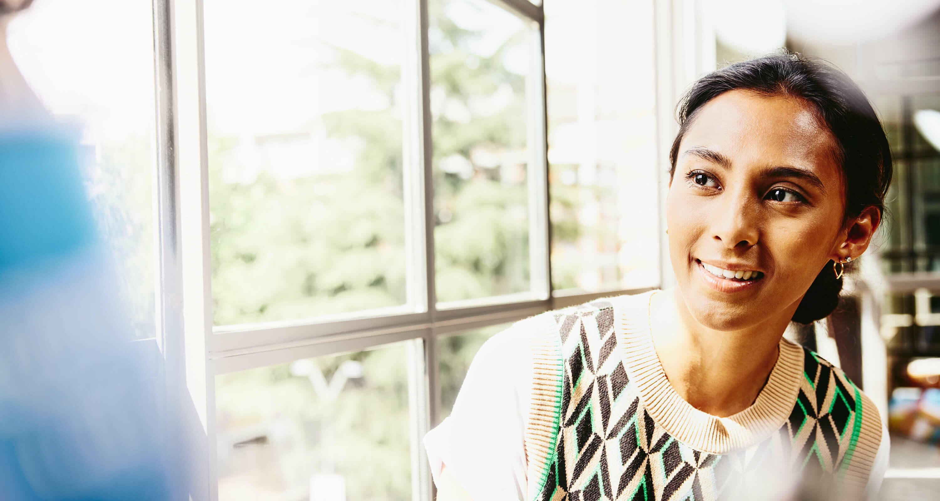 a woman in a colourful top seated by the window while talking to her colleague