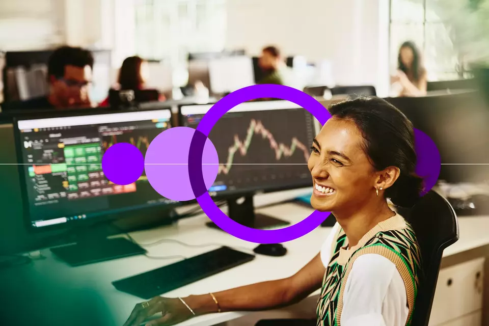 an image of a woman smiling while sitting behind her desk, screens displaying financial information.