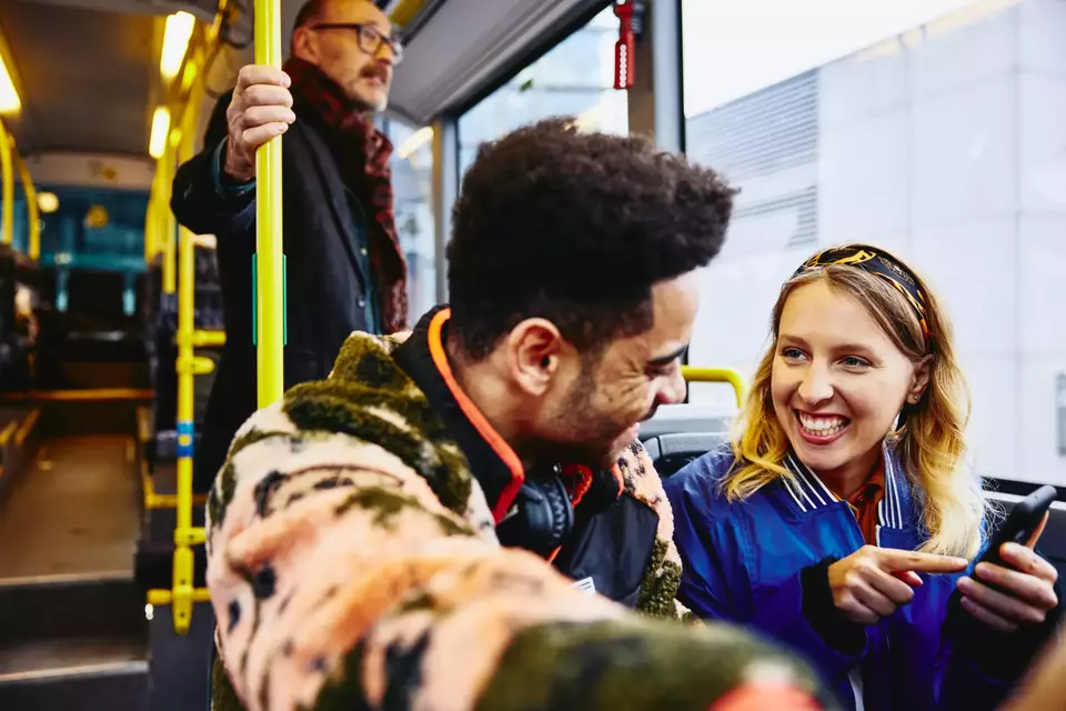 Womale and boy having a conversation, smiling, sitting in a bus. Looking at a phone. Other people in the background.
