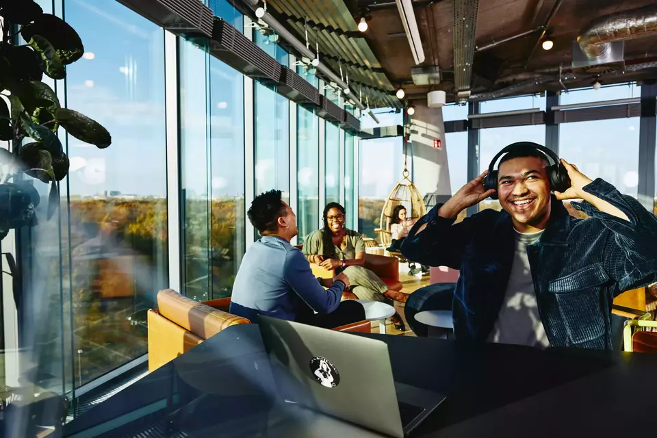 Smiling male with headphones and laptop sitting in a lounge environment.
