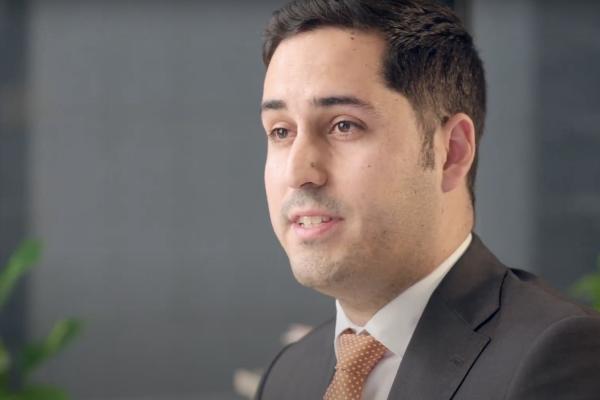 man with brown hair, in a suit with orange tie talking to the camera